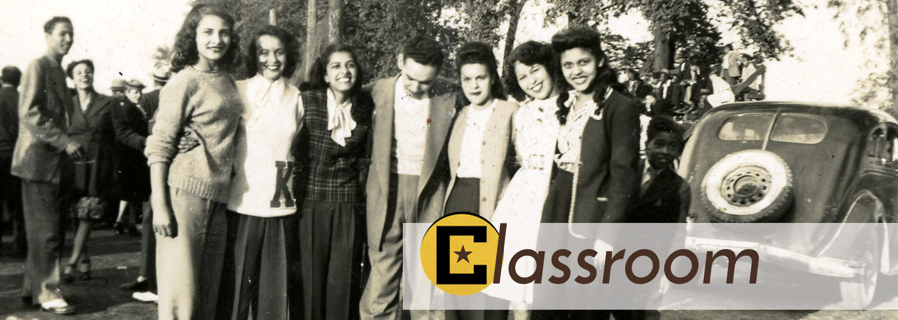 Picture of 7 young people standing together outside with arms linked. Old car in background.
