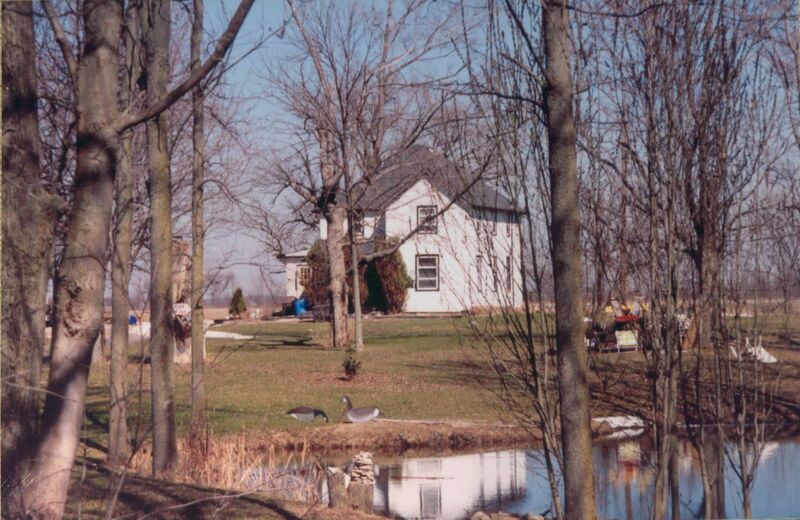 A medium sized white house with a grey roof stands in the background with a lawn and some trees standing in front and a river in the foreground. The photo was taken in winter, and the trees all have no leaves. 