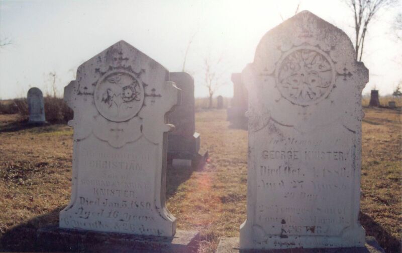 Two white gravestones with circular decorative carvings at their top back lit in a cemetery. The writing is faded and they look weathered. 