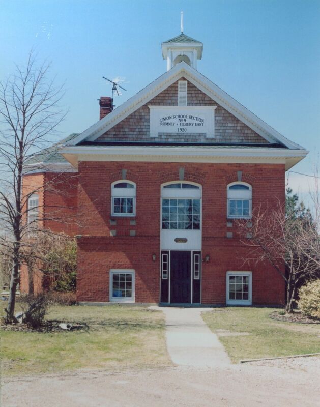 Photo of a red brick building, with an angled grey roof and a small white bell tower without the bell. There are trees on the sides of the building and lawn, with their leaves missing for winter. The sign at the top of the building's front facade reads "Union School Section No 9 Romney-Tilbury East 1920," and was an original part of the structure when it was a schoolhouse. 
