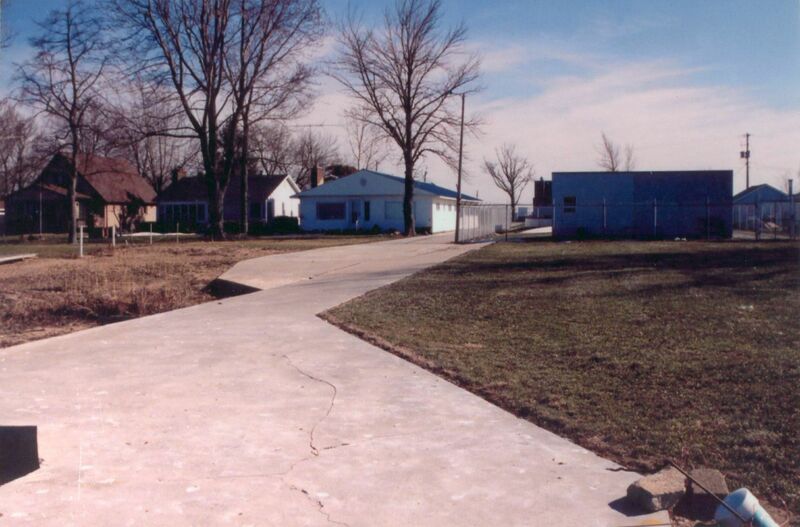 Photo of a concrete pathway in the foreground between grass. Houses are visible on the left side of the  path in the distance, behind a few trees. On the right side there is a back of a rectangular pumping station. 