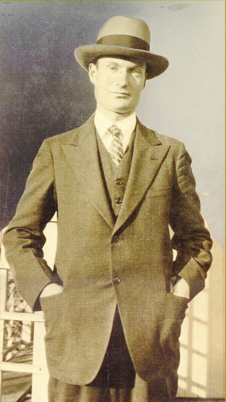Raymond Knister standing looking directly at the camera with a bowler hat, a three piece suit and his hands in his suit pockets. 