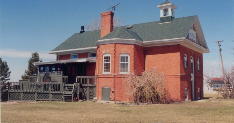 Side of a large rectangular red brick building with a grey roof. There is a curved room protruding from the building face, and a wooden porch at the back. The lawn is brown and the tree beside the curved wall has no leaves. 