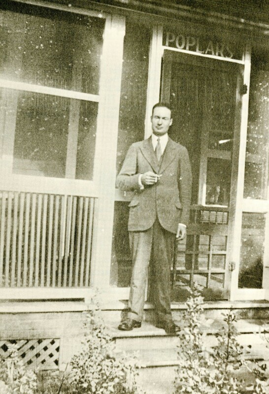 Raymond Knister is standing holding a cigarette looking directly at the camera on the second step of three, in front of a wooden porch. There is a sign which says "Poplars" above the door to the porch. The whole photo is a faded sepia tone. 
