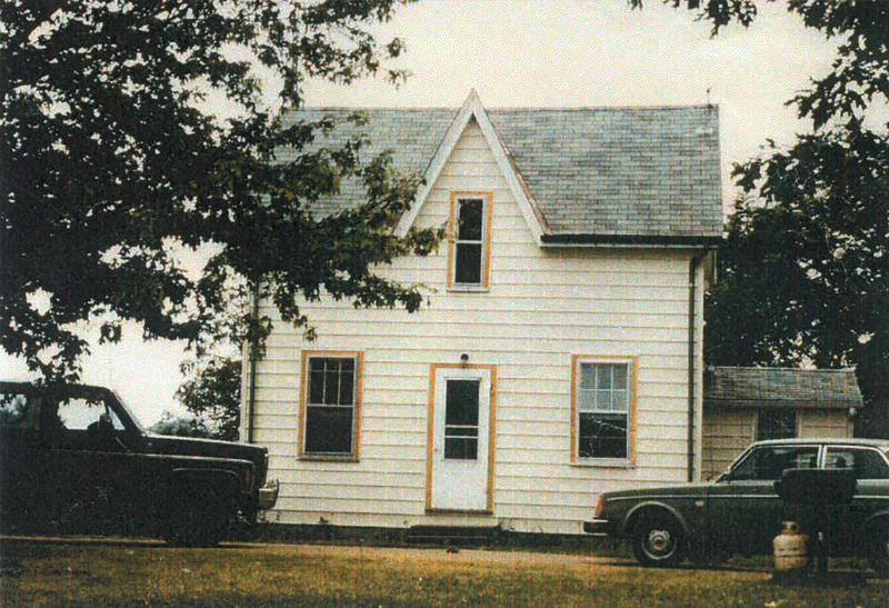 A small white panelled house with a grey roof and wood details around the door and three windows, with two cars parked in front. The cars look like they're from the early 1970s and are visible on a side profile. 