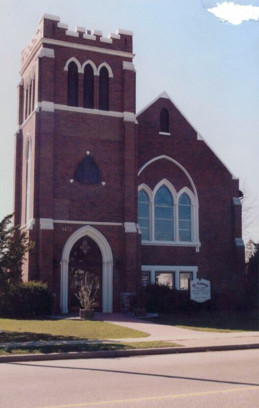 A photo of a brick church with a rectangular tower on the left side of the building, and a shorter right side, with an inverted triangular roof. 