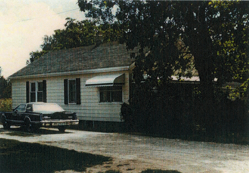 A small white panelled house with a grey roof. There's a black car parked in front on the driveway, which looks to be from the 1980s. 