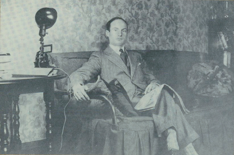 Raymond Knister sitting on a couch with his legs crossed looking at the camera with a newspaper in his lap. There is floral wallpaper and a lamp pointed at the couch from a side table.