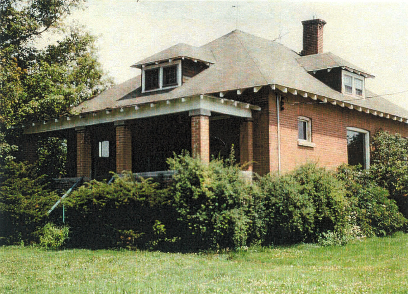 Photo of a small red brick bungalow, with a small lawn and bushes surrounding its outer walls. 