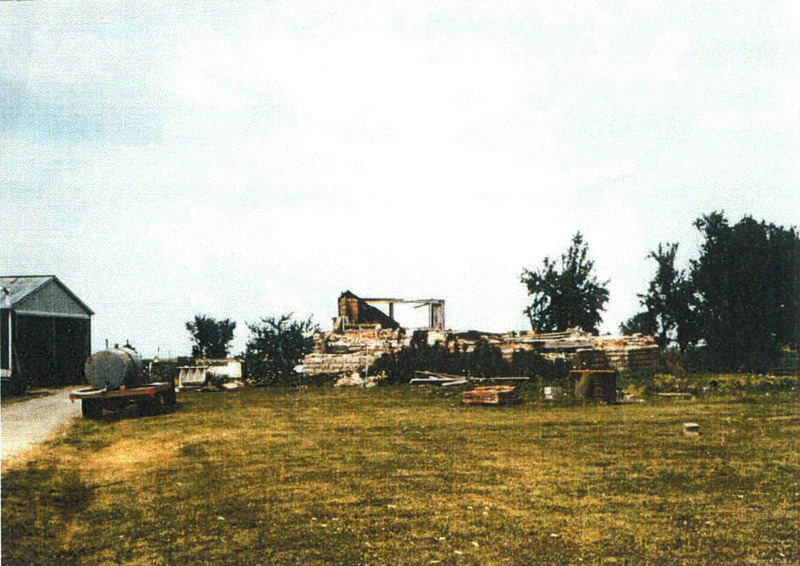 Photo of a grassy area and what appears to be the remains of a broken down house, with a small truck on the side and a small building partially visible on the left side. 