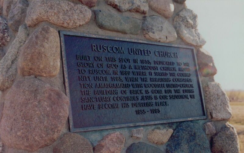 Photo of a black metal sign inlaid on stone wall. Title reads: "Ruscom United Church." Text reads "Built on this spot in 1855, dedicated to the glory of God as a Methodist Church. Moved to Ruscom in 1889 where it served the community until 1985, where the remaining congregations amalgamated with Woodslee United Church. The building of brick is gone but the living sanctuary continues. Jesus is our salvation, we have become his dwelling place. 1855-1985."