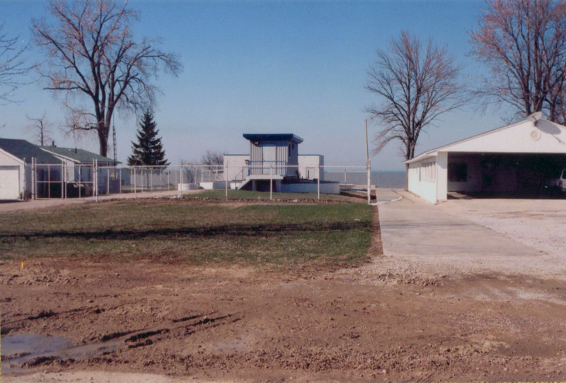 A photo of a fenced grey pumping station on the left, a narrow concrete pathway in between it and a garage of a house and its driveway. The foreground is a muddy path across the bottom of the photo. 