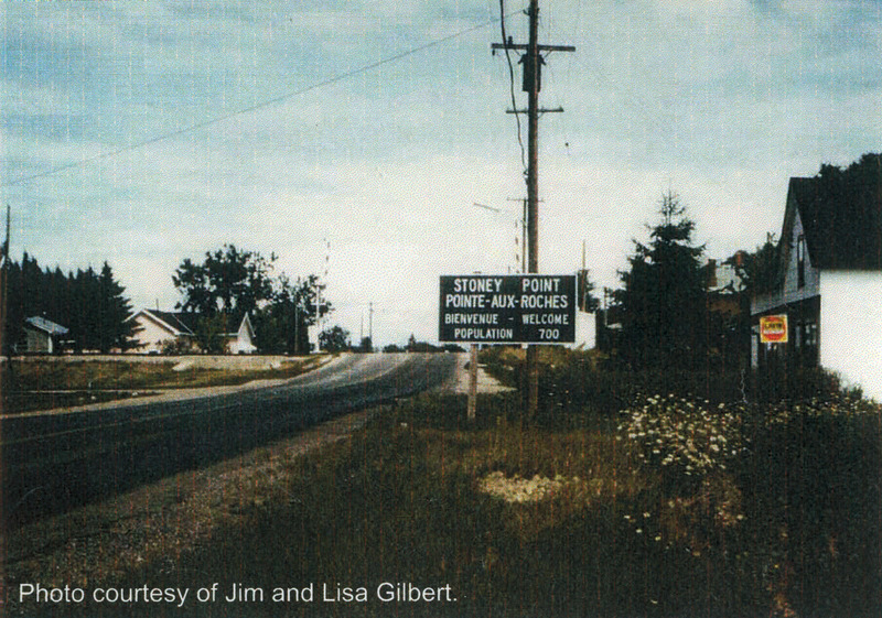 Photo of the Welcome to Stoney Point highway sign with Highway 7 running beside it. There are some houses on wither side of the highway and a telephone pole behind the road sign. 