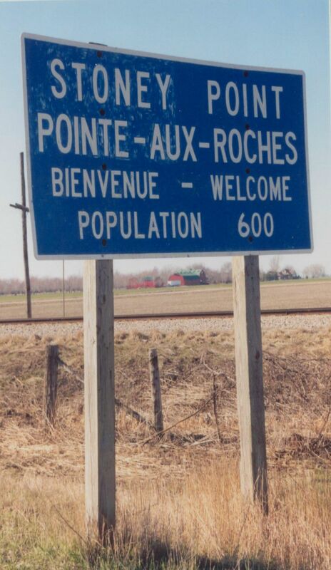 Blue and white highway sign on wooden stakes. The sign reads: "Stoney Point, Pointe-Aux-Roches, Bienvenue - Welcome, Population 600."
