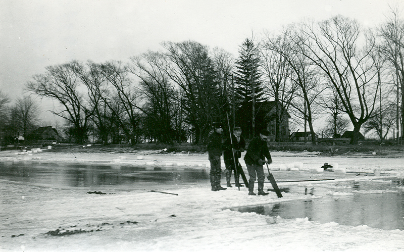 Black and white photo of three men standing on a frozen river with various ice harvesting equipment in their hands. 