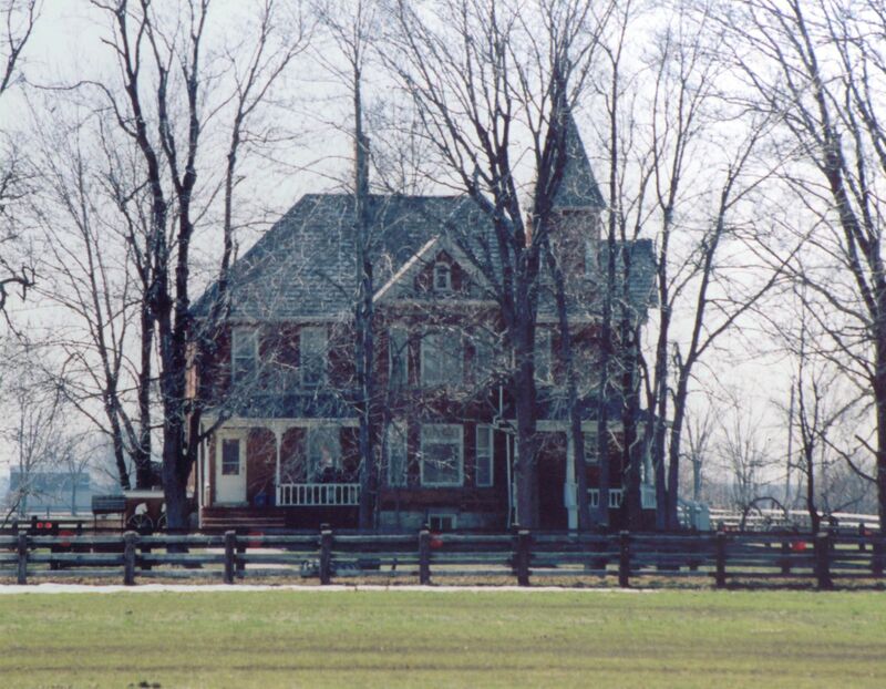 A large house of red brick and a grey roof stands in the background with trees, a fence and a lawn in front. The house has a small circular tower jutting from the right side and white detailing. 