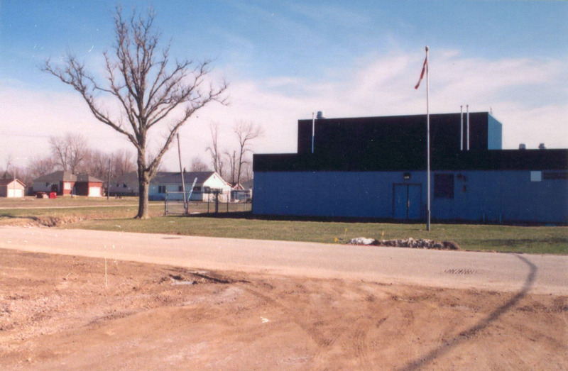 A large rectangular building with two levels in black and dark grey. The lower rectangle is larger with a smaller black rectangular floor on top. There is a flag pole with a Canadian flag in front, a narrow road running horizontally and a section of dirt in the foreground. 