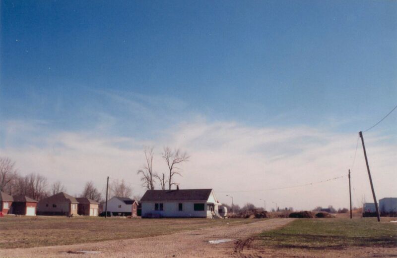 A photo of four white and beige cottages in the distance under a large blue sky. There is a dirt road running beside them and a patch of grass on the far left side beside the road. 