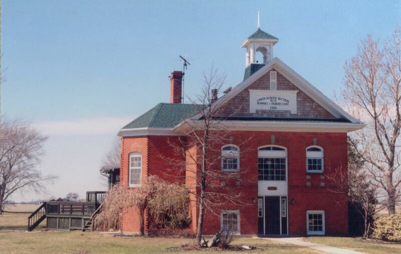 Photo of a red brick building, with an angled grey roof and a small white bell tower without the bell. There is a wooden porch visible on the left side, at the back of the building. There are 4-5 trees on the sides of the building and lawn, with their leaves missing for winter.   The sign at the top of the building's front facade reads "Union School Section No 9 Romney-Tilbury East 1920," and was an original part of the structure when it was a schoolhouse. 