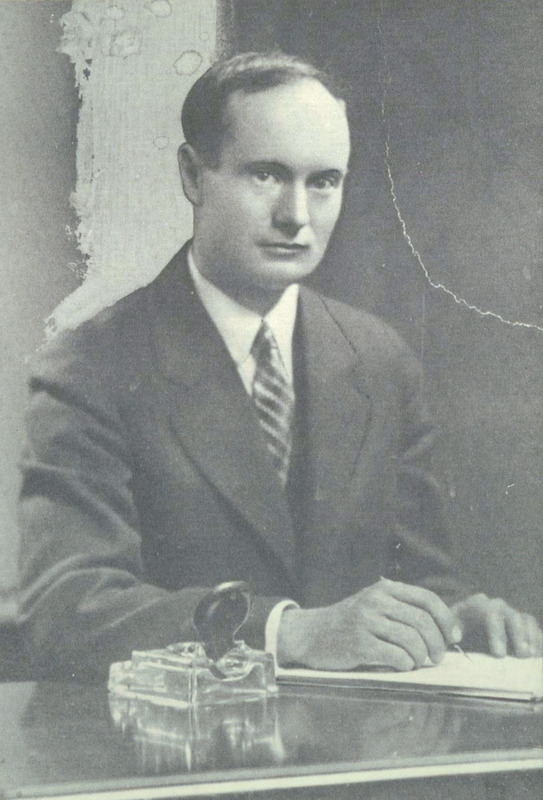 Black and white photo of Raymond Knister looking at the camera with a pen in his hands and paper. He's seated at a desk and wearing a suit with a striped tie. 