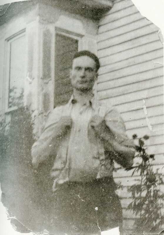 A faded black and white photo with the top right and bottom left corners ripped. Raymond Knister is standing in the middle in front of a house and a few small plants, staring forward. He has short dark hair, his hands are holding his suspenders. 