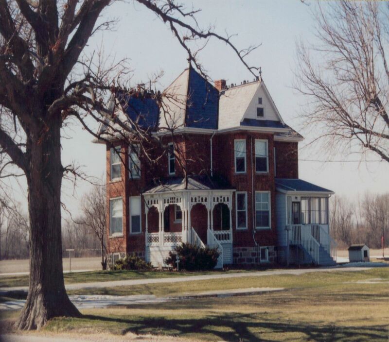 A photo of a red brick house with white detailing and grey roof surrounded by a yellowing lawn. The spine of the roof is covered in metal spikes. There is a tree with no leaves to the side in the foreground. 