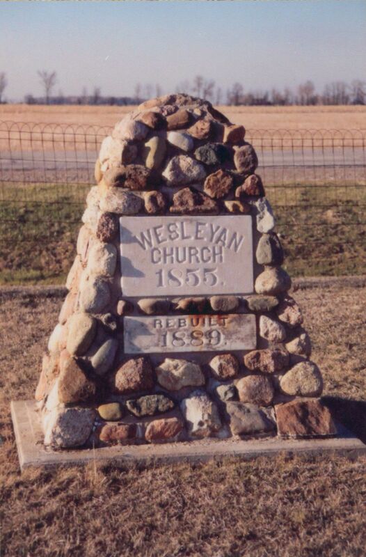 Photo of 2 white stone sign inlaid on to short stone pillar in front of a fence. The larger top sign reads "Wesleyan Church 1855" and the lower smaller one reads "Rebuilt 1889."