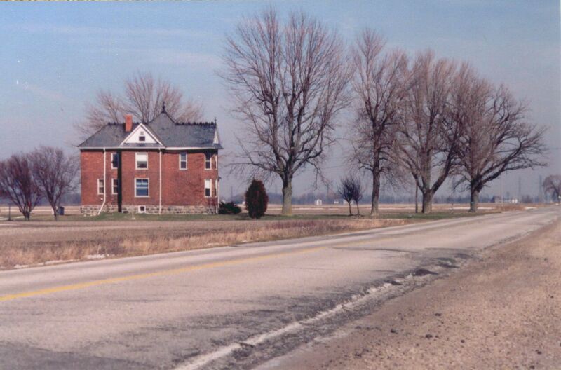 A  red brick house with a grey roof stands to the side of a small two lane road. There are trees without their leaves lining the road side beside the house. 