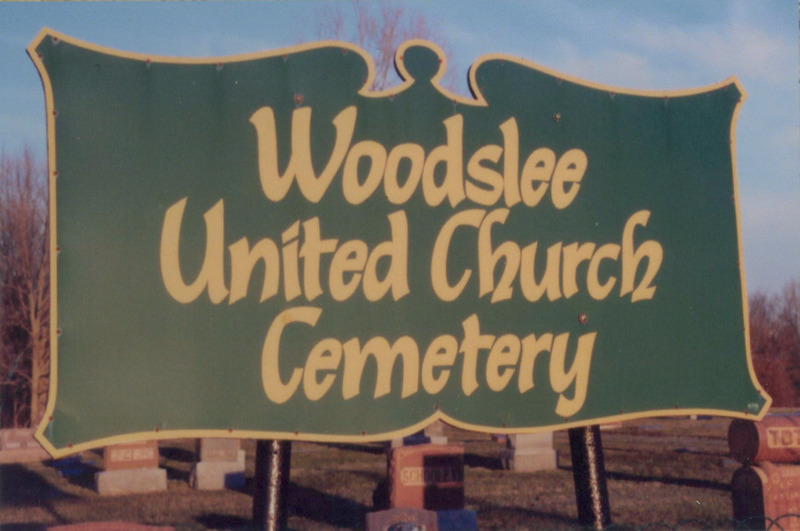 A dark green sign with a yellow border and yellow text stating: "Woodslee United Church Cemetery." 