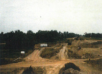 A gravel pit with three dirt roads, two parallel running vertically and one perpendicular running horizontally through them both. There's trees in the middle distance and a white sky. 