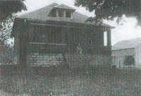Black and white photo of a small bungalow with a triangular roof, and a porch. There's a child sitting at the top of the steps. 