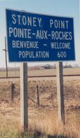 Blue and white highway sign on wooden stakes. The sign reads: "Stoney Point, Pointe-Aux-Roches, Bienvenue - Welcome, Population 600."