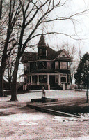 Sepia tinted photo of a large house with a sprawling lawn. The house has a curving porch and a small circular spire on one side. There's a statue of a dog on the lawn, and some snow on the ground. The trees lining the lawn have no leaves. 